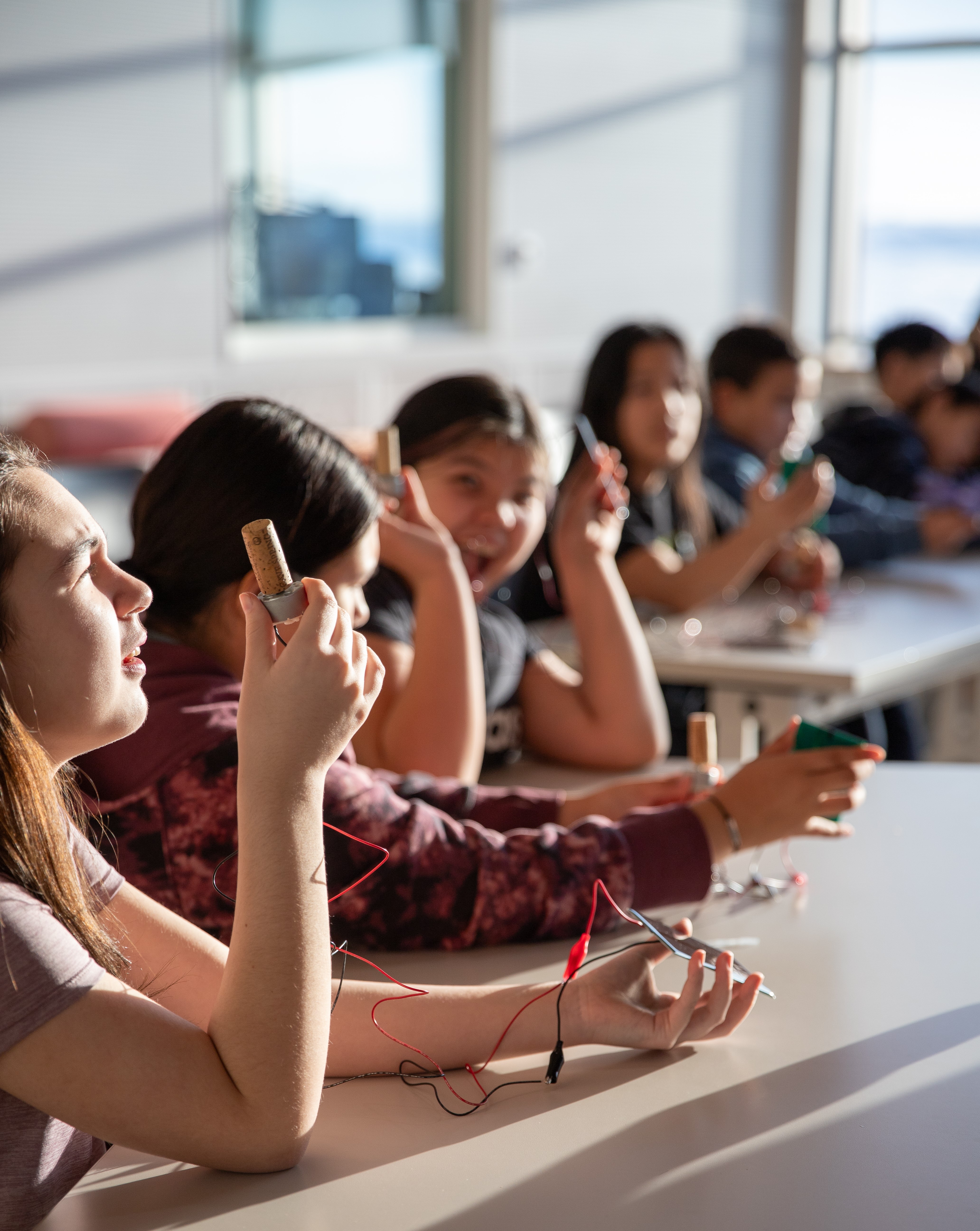 Young teenagers sit in a row of desks, each holds a small solar panel connected to a tiny fan.